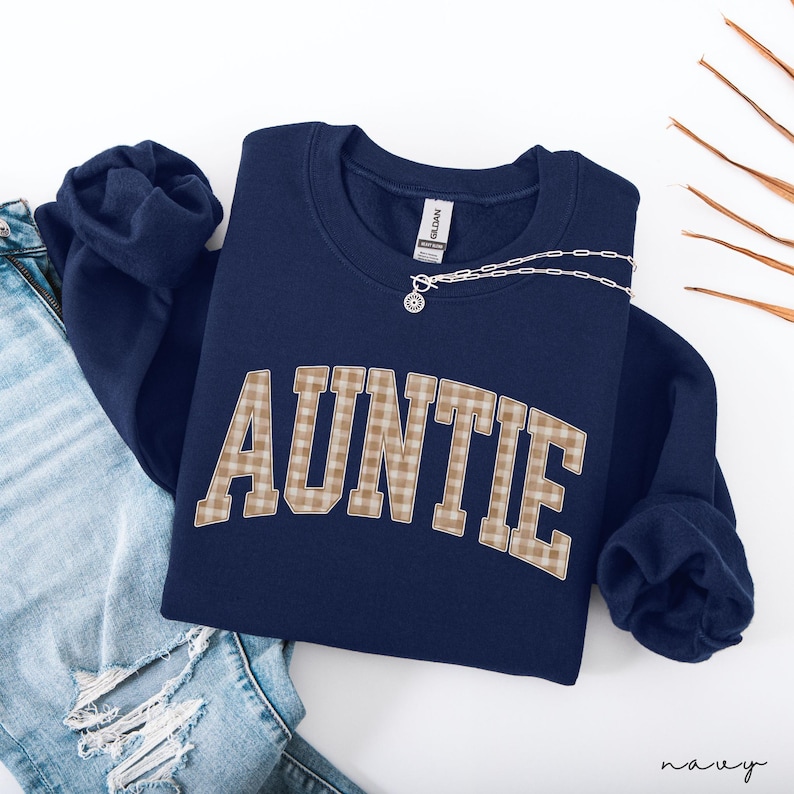 May include: Navy blue sweatshirt with the word "AUNTIE" in a plaid pattern across the front. A silver necklace with a pendant is draped over the sweatshirt. The sweatshirt is folded next to a pair of blue jeans.