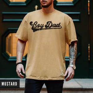 Puede incluir: Camiseta color mostaza con la frase "Boy Dad" en negro desgastado. La camiseta tiene mangas cortas y cuello redondo. La imagen muestra la camiseta en una persona.