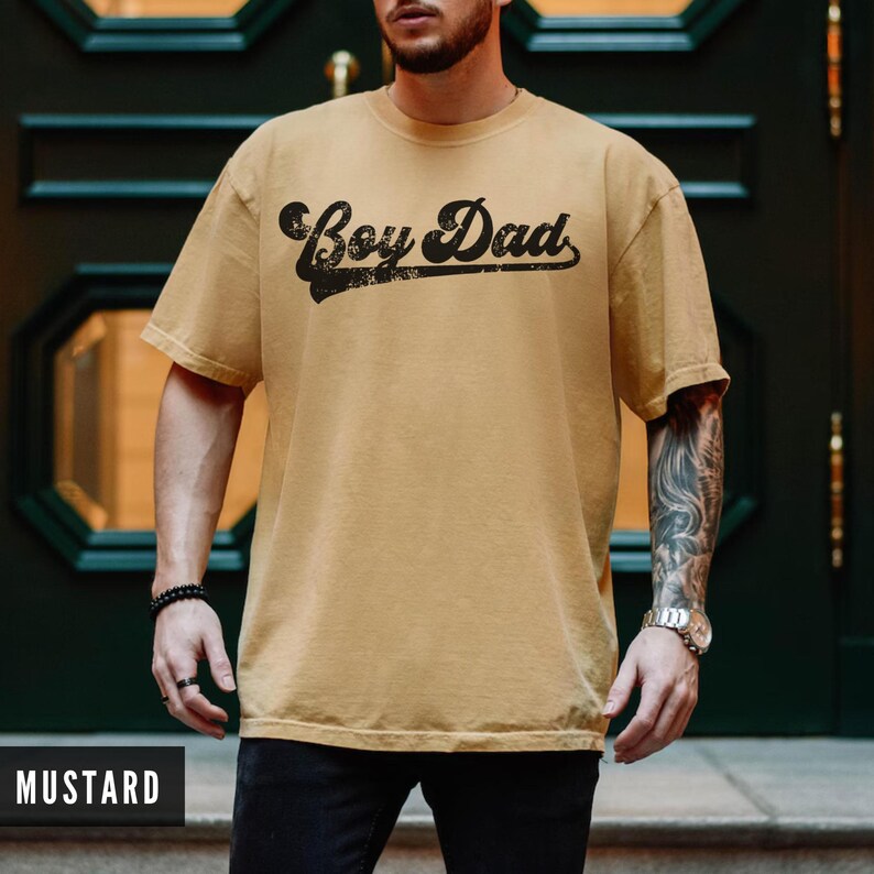 Puede incluir: Camiseta color mostaza con la frase "Boy Dad" en negro desgastado. La camiseta tiene mangas cortas y cuello redondo. La imagen muestra la camiseta en una persona.