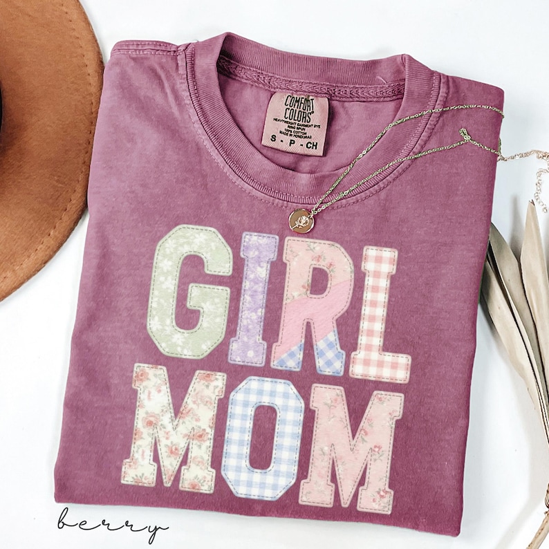May include: Berry-colored t-shirt with the words "GIRL MOM" in patchwork letters. The letters are in various floral and gingham patterns. A delicate gold necklace with a pendant rests on the shirt. The shirt has a crew neck and a tag that says "Comfort Colors".