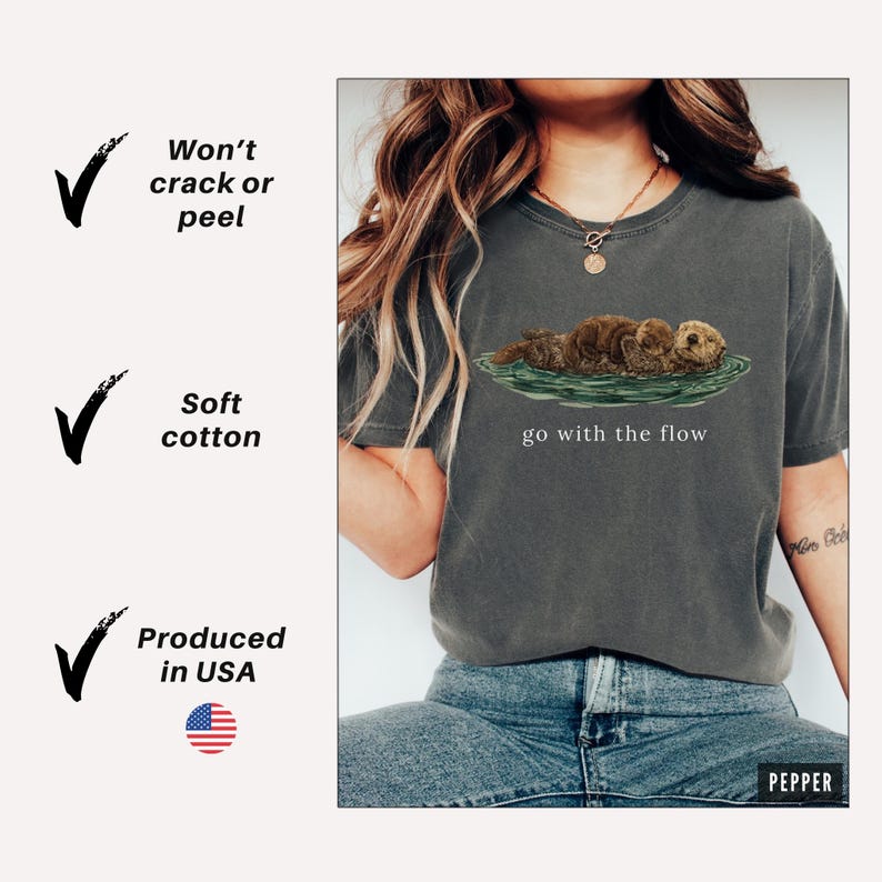 May include: Dark grey t-shirt with an illustration of two otters in water and the text "go with the flow". The shirt is made of soft cotton. The t-shirt is produced in the USA.