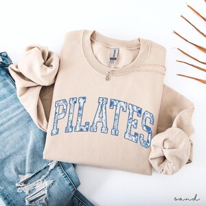 May include: A sand-colored sweatshirt with the word "PILATES" in blue floral lettering. A silver necklace with a small pendant rests on the sweatshirt. The sweatshirt is paired with light-wash denim jeans.