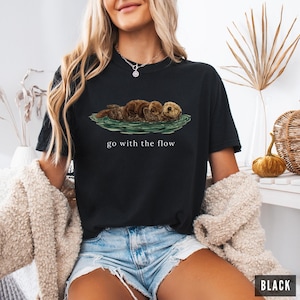 May include: Black crew neck t-shirt with an illustration of three otters on water. The words "go with the flow" are printed in white beneath the otters. The shirt is a casual style.