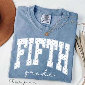 May include: A blue jean coloured t-shirt with a crew neckline and a Comfort Colors label. The front features the word FIFTH in large white block letters with a grey polka dot pattern, and the word grade in white cursive script below it.