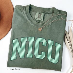 May include: A moss green short-sleeve t-shirt with a crew neckline and the text NICU printed in large, light green and dark green vertical striped block letters across the chest. The shirt features a Comfort Colors brand label at the neck.