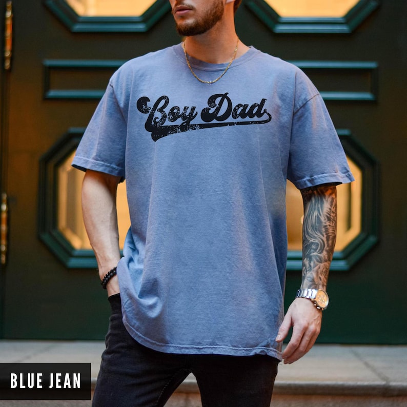 Puede incluir: Camiseta azul vaquero con el texto "Boy Dad" en fuente negra. La camiseta tiene mangas cortas y un corte holgado. La imagen muestra la camiseta en una persona.