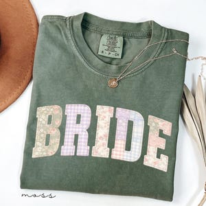 Może przedstawiać: Zielona koszulka w kolorze mchu z napisem "BRIDE" w patchworkowym wzorze. Patchwork zawiera kwiatowe i kratowane wzory w kolorze różowym, fioletowym i niebieskim. Złoty naszyjnik z wisiorkiem spoczywa na koszulce. Koszulka jest złożona.