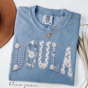 May include: A blue jean Comfort Colors t-shirt with the word "DOULA" appliqued in white, blue gingham, and floral patterns. Each letter is adorned with a small daisy. A gold necklace with a pendant rests on the shirt.