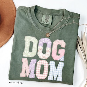 May include: A moss green t-shirt with the words "DOG MOM" in colourful, patchwork-style letters. The shirt has a crew neck and short sleeves. A gold necklace with a pendant rests on the shirt. A brown hat is partially visible in the upper left corner.
