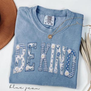 Puede incluir: Una camiseta azul Comfort Colors con las palabras "BE KIND" aplicadas en un diseño de patchwork de patrones florales y de cuadros. Un collar de oro con un colgante descansa sobre la camisa. La camisa está etiquetada como "blue jean".