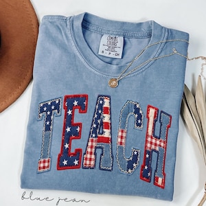 Op de afbeelding: Een blauw Comfort Colors T-shirt met het woord "TEACH" op de voorkant in een patriottisch ontwerp. De letters zijn gemaakt van denim, rood geruit en witte sterren. Een gouden ketting met een hanger ligt over het shirt.