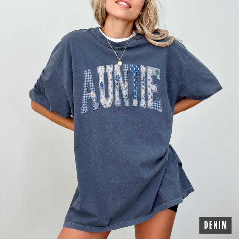 May include: Auntie graphic tee in denim blue. The word "AUNTIE" is appliqued across the front in a patchwork design with floral and gingham patterns. The shirt has a relaxed fit and short sleeves. The word "DENIM" is printed in a rectangle in the bottom right corner.
