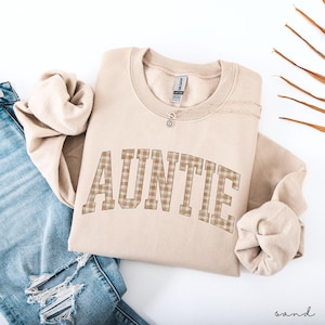 May include: A sand-colored sweatshirt with the word "AUNTIE" in a plaid pattern across the front. A delicate necklace with a small pendant rests on the sweatshirt. The sweatshirt is folded next to a pair of blue jeans.