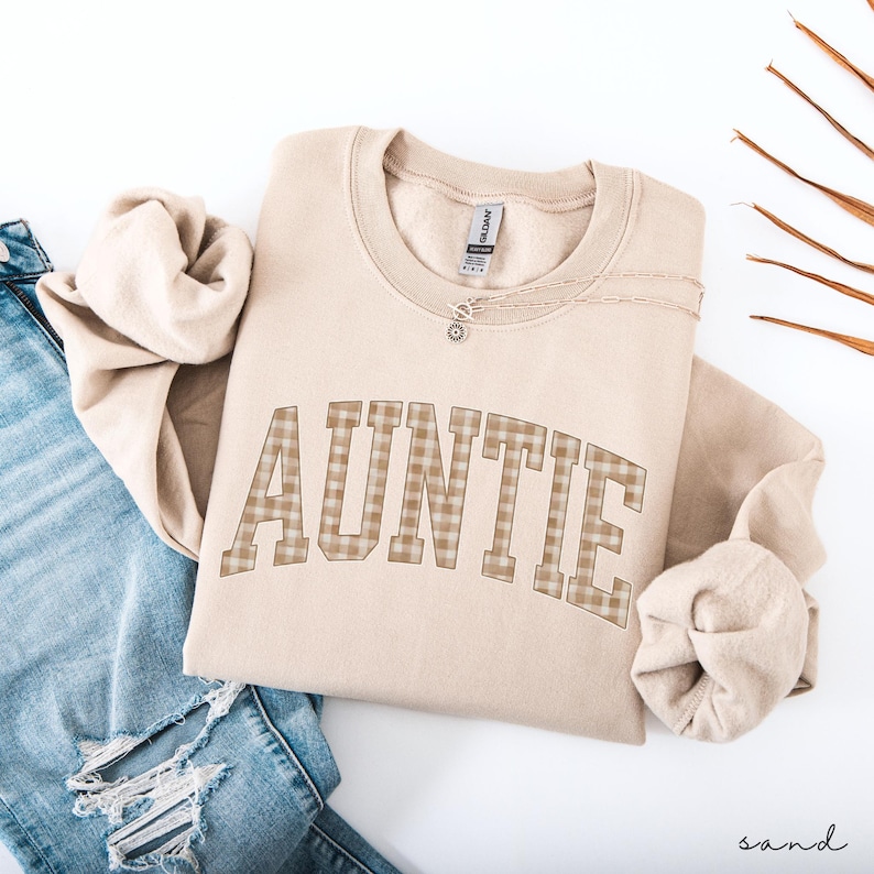 May include: A sand-colored sweatshirt with the word "AUNTIE" in a plaid pattern across the front. A delicate necklace with a small pendant rests on the sweatshirt. The sweatshirt is folded next to a pair of blue jeans.