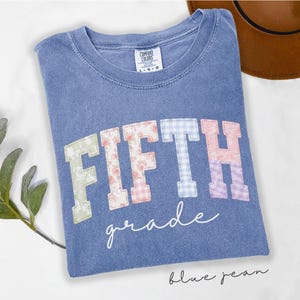 Puede incluir: Una camiseta azul de cuello redondo con la palabra FIFTH en letras grandes de bloque con patrones multicolores y la palabra grade en cursiva blanca. La prenda incluye una etiqueta Comfort Colors de talla L y un acabado de tela desgastada.