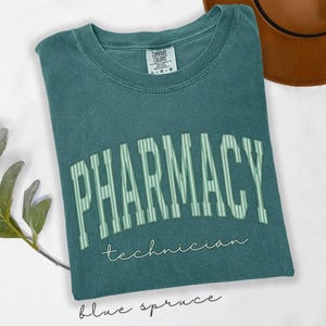 K&ouml;nnte beinhalten: Ein blaugr&uuml;nes T-Shirt mit Rundhalsausschnitt und verwaschenem Vintage-Finish. Auf der Brust steht das Wort PHARMACY in gro&szlig;en, gebogenen, hellgr&uuml;n gestreiften Blockbuchstaben, darunter das Wort technician in kleiner, wei&szlig;er Schreibschrift.