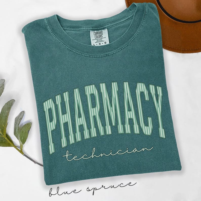K&ouml;nnte beinhalten: Ein blaugr&uuml;nes T-Shirt mit Rundhalsausschnitt und verwaschenem Vintage-Finish. Auf der Brust steht das Wort PHARMACY in gro&szlig;en, gebogenen, hellgr&uuml;n gestreiften Blockbuchstaben, darunter das Wort technician in kleiner, wei&szlig;er Schreibschrift.