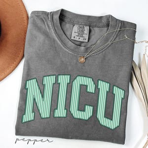 May include: A charcoal gray short-sleeve t-shirt with a crew neckline. The shirt features the text NICU in large, block-style letters with a light green and white vertical striped pattern. A small silver-toned necklace with a circular pendant rests on the collar.
