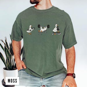 May include: Green t-shirt featuring a cartoon of three ducks exercising with weights. The ducks are lifting dumbbells, bench pressing, and using a step. The word "MOSS" is printed on a white rectangle at the bottom.
