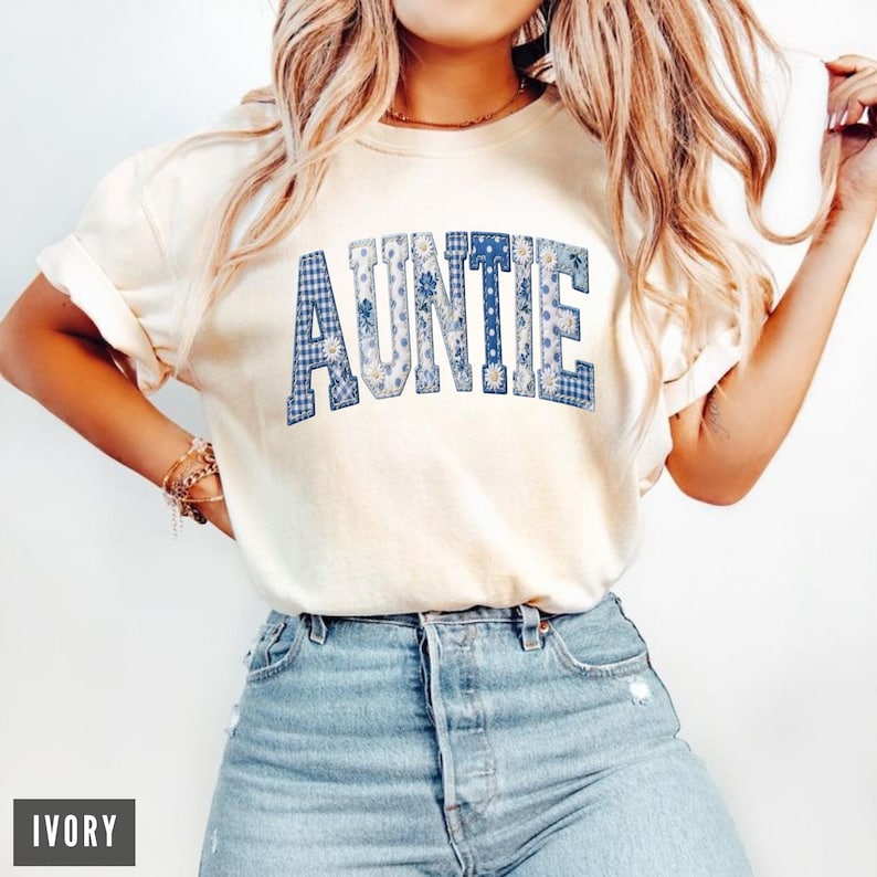 May include: Ivory-colored t-shirt with the word "AUNTIE" in blue letters. The letters are filled with a mix of floral and gingham patterns. The shirt is paired with light-wash denim jeans.