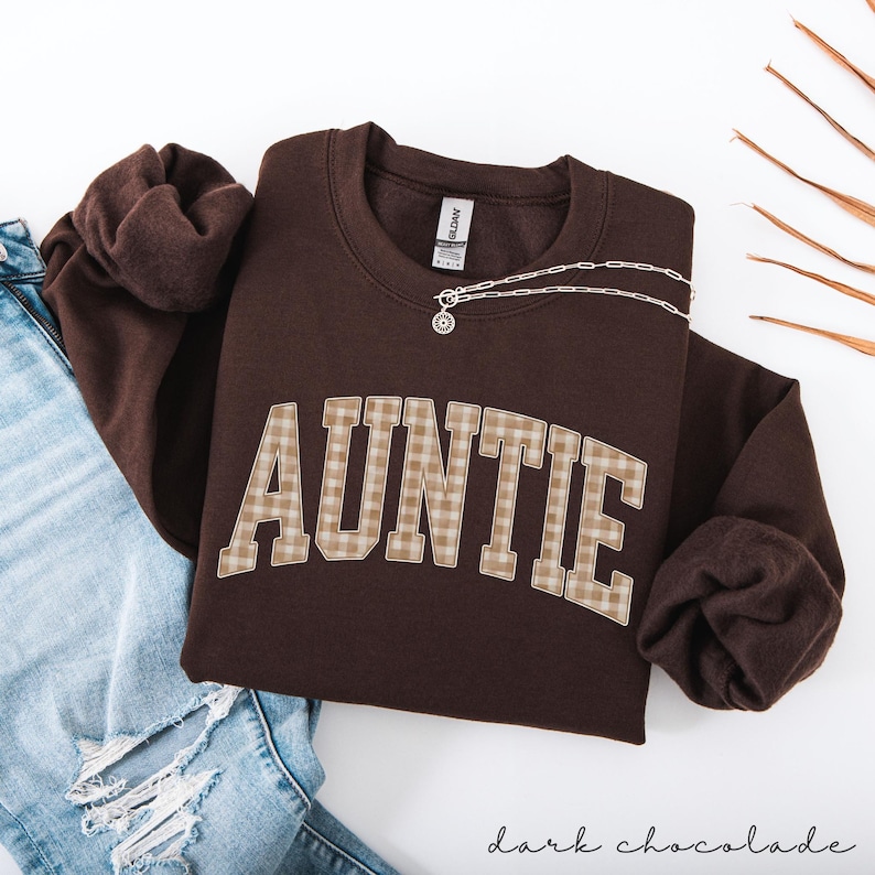 May include: A dark chocolate-colored sweatshirt with the word "AUNTIE" in a plaid design. A silver necklace with a pendant rests on the sweatshirt. The sweatshirt is folded next to a pair of blue jeans.