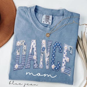 Könnte beinhalten: Ein blaues Comfort Colors T-Shirt mit dem Wort "DANCE", das in verschiedenen blauen Blumen- und Tupfenmustern appliziert ist. Das Wort "Mom" ist darunter in Schreibschrift geschrieben. Eine goldene Halskette liegt auf dem Shirt.