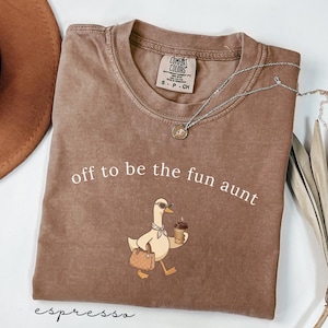 May include: A brown short-sleeved t-shirt with a crew neckline and a graphic print of a duck wearing sunglasses and a scarf, holding a coffee cup and a handbag. The text "off to be the fun aunt" is printed above the duck in white lettering.