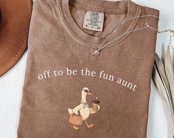 May include: A brown short-sleeved t-shirt with a crew neckline and a graphic print of a duck wearing sunglasses and a scarf, holding a coffee cup and a handbag. The text "off to be the fun aunt" is printed above the duck in white lettering.