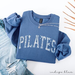 May include: Indigo blue sweatshirt with the word "PILATES" in a floral design across the front. A silver necklace with a pendant is on the sweatshirt. The sweatshirt is folded on top of a pair of blue jeans.