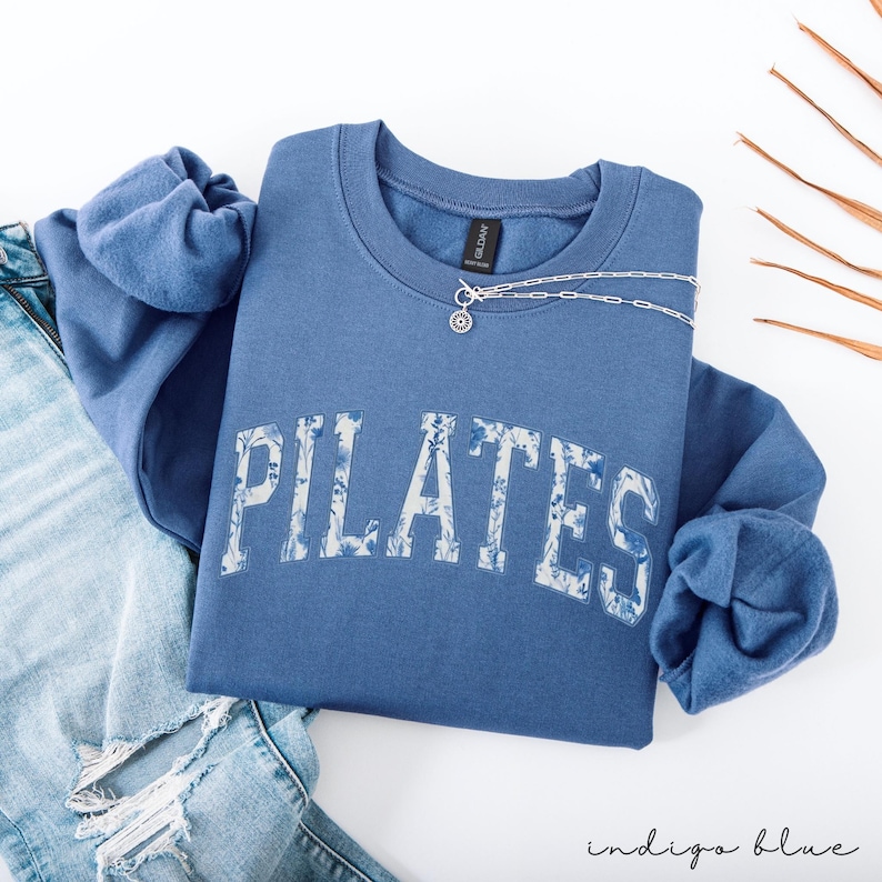 May include: Indigo blue sweatshirt with the word "PILATES" in a floral design across the front. A silver necklace with a pendant is on the sweatshirt. The sweatshirt is folded on top of a pair of blue jeans.