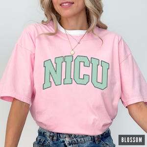 May include: A light pink short-sleeve t-shirt featuring the text NICU in large, arched, green and white vertical striped block letters. The shirt has a crew neckline and is worn with a gold pendant necklace and blue denim jeans.