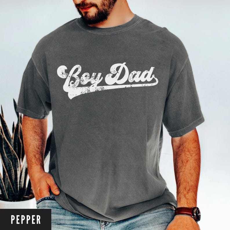 Puede incluir: Camiseta gris con la frase "Boy Dad" en una fuente blanca desgastada. La camiseta es de color liso y tiene mangas cortas. La palabra "Pepper" est&aacute; en un rect&aacute;ngulo negro en la parte inferior.