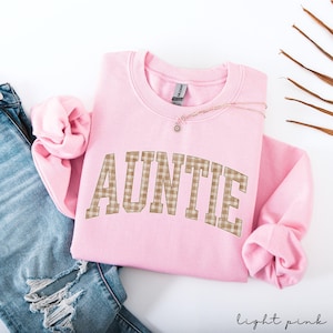 May include: A light pink sweatshirt with the word "AUNTIE" in a brown plaid design across the front. A delicate necklace with a small pendant rests on the sweatshirt. The bottom right corner of the image has the words "light pink".