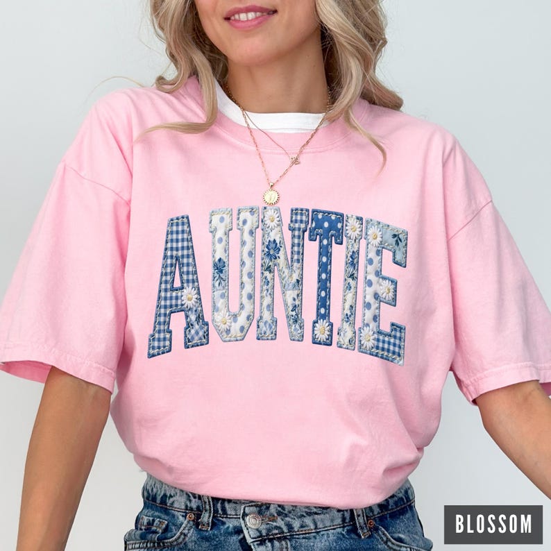 May include: A pink t-shirt with the word "AUNTIE" in a patchwork design. The letters are a mix of blue gingham, floral, and solid blue fabrics. The shirt has a white collar and is paired with blue jeans.