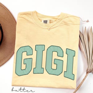 Pode incluir: Uma t-shirt de manga curta amarelo-manteiga com decote redondo e a palavra GIGI impressa em letras grandes de bloco verdes com riscas verticais no peito. A t-shirt apresenta uma etiqueta da marca Comfort Colors na zona do pescoço.