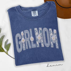 May include: A blue denim t-shirt with the words "GIRL MOM" in a white and blue floral and gingham pattern. The shirt has a crew neck and the text "denim" is written in cursive at the bottom.