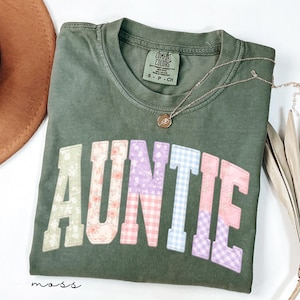 May include: An olive green t-shirt with the word "AUNTIE" in colorful patchwork letters. The shirt has a crew neck and a small tag at the neckline. A gold necklace with a pendant rests on the shirt.