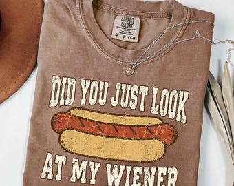 May include: A brown short-sleeve t-shirt featuring a graphic of a hot dog and the text "DID YOU JUST LOOK AT MY WIENER" in a distressed white font. The shirt has a Comfort Colors brand label at the neck and is made of 100% cotton fabric.