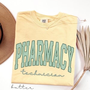K&ouml;nnte beinhalten: Ein buttergelbes Kurzarm-T-Shirt mit dem Wort PHARMACY in gro&szlig;en, gebogenen, gr&uuml;n gestreiften Blockbuchstaben und dem Wort technician darunter in gr&uuml;ner Schreibschrift. Das T-Shirt hat ein Nackenetikett der Marke Comfort Colors.