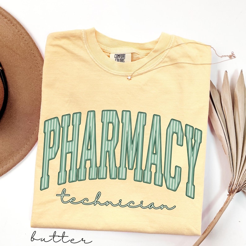 K&ouml;nnte beinhalten: Ein buttergelbes Kurzarm-T-Shirt mit dem Wort PHARMACY in gro&szlig;en, gebogenen, gr&uuml;n gestreiften Blockbuchstaben und dem Wort technician darunter in gr&uuml;ner Schreibschrift. Das T-Shirt hat ein Nackenetikett der Marke Comfort Colors.
