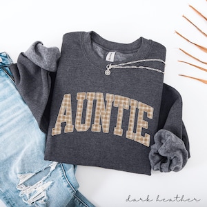 May include: Dark heather gray sweatshirt with the word "AUNTIE" in a plaid design. A silver necklace with a pendant rests on the sweatshirt. The sleeves are cuffed, and the bottom of the sweatshirt is visible. The words "dark heather" are written in script at the bottom.