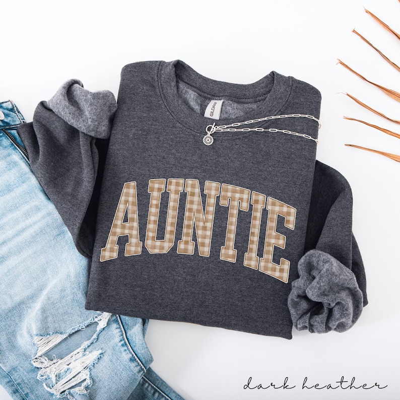 May include: Dark heather gray sweatshirt with the word "AUNTIE" in a plaid design. A silver necklace with a pendant rests on the sweatshirt. The sleeves are cuffed, and the bottom of the sweatshirt is visible. The words "dark heather" are written in script at the bottom.