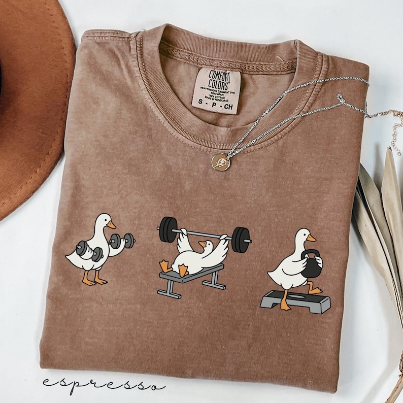 May include: Espresso-coloured Comfort Colors t-shirt with cartoon geese lifting weights. The geese are shown doing gym exercises with dumbbells, bench press, and kettlebell. The shirt has a small tag that says "S-P-CH". A silver necklace with a pendant is also visible.