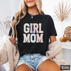 May include: Black t-shirt with the words "GIRL MOM" in pastel and patterned letters. The shirt is a crew neck and is being worn by a person. The word "BLACK" is in a black rectangle in the bottom right corner.