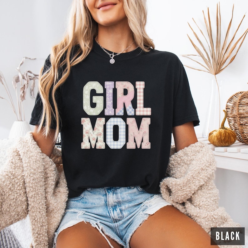 May include: Black t-shirt with the words "GIRL MOM" in pastel and patterned letters. The shirt is a crew neck and is being worn by a person. The word "BLACK" is in a black rectangle in the bottom right corner.