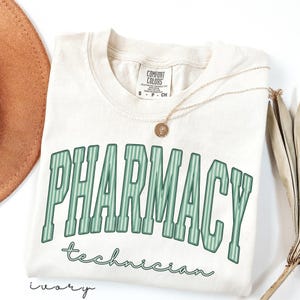 K&ouml;nnte beinhalten: Ein elfenbeinfarbenes T-Shirt mit dem Wort PHARMACY in gro&szlig;en, gr&uuml;n gestreiften Blockbuchstaben im College-Stil, darunter steht das Wort technician in gr&uuml;ner Schreibschrift. Eine goldfarbene Halskette mit kleinem runden Anh&auml;nger liegt auf dem Shirt.