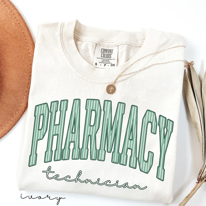 K&ouml;nnte beinhalten: Ein elfenbeinfarbenes T-Shirt mit dem Wort PHARMACY in gro&szlig;en, gr&uuml;n gestreiften Blockbuchstaben im College-Stil, darunter steht das Wort technician in gr&uuml;ner Schreibschrift. Eine goldfarbene Halskette mit kleinem runden Anh&auml;nger liegt auf dem Shirt.