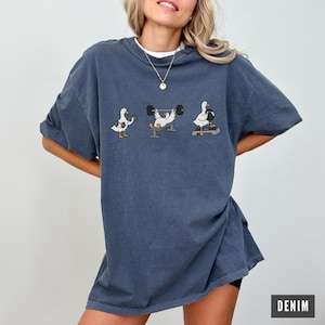May include: Denim blue t-shirt with a comical design of three geese weightlifting. The geese are shown in different weightlifting positions, using barbells and dumbbells. The word "DENIM" is printed in white on a dark grey rectangle.