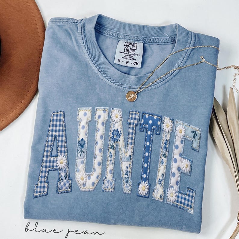 May include: A blue jean Comfort Colors t-shirt with the word "AUNTIE" in a patchwork design. The letters feature gingham, floral, and polka dot patterns. A gold necklace with a pendant is draped across the shirt.