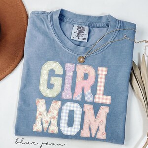May include: Blue jean colored t-shirt with the words "GIRL MOM" in patchwork letters. The letters are in various pastel floral and gingham patterns. A gold necklace with a pendant is draped across the shirt.